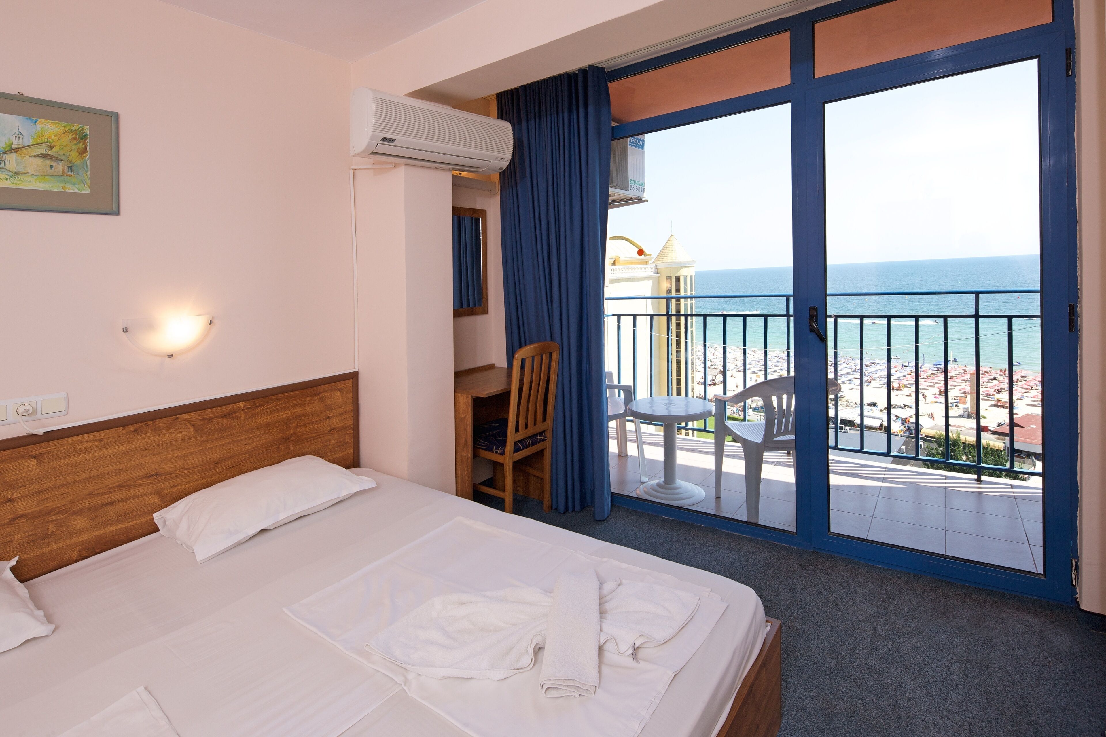 A hotel room with a bed, a desk, a chair, and a balcony with a view of the beach and ocean.