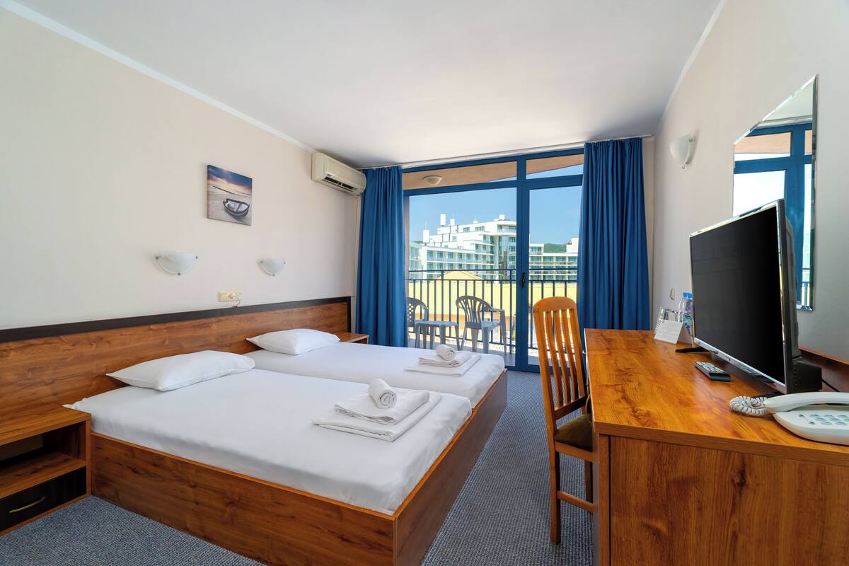 A hotel room with two beds, a desk with a TV, and a balcony with chairs.