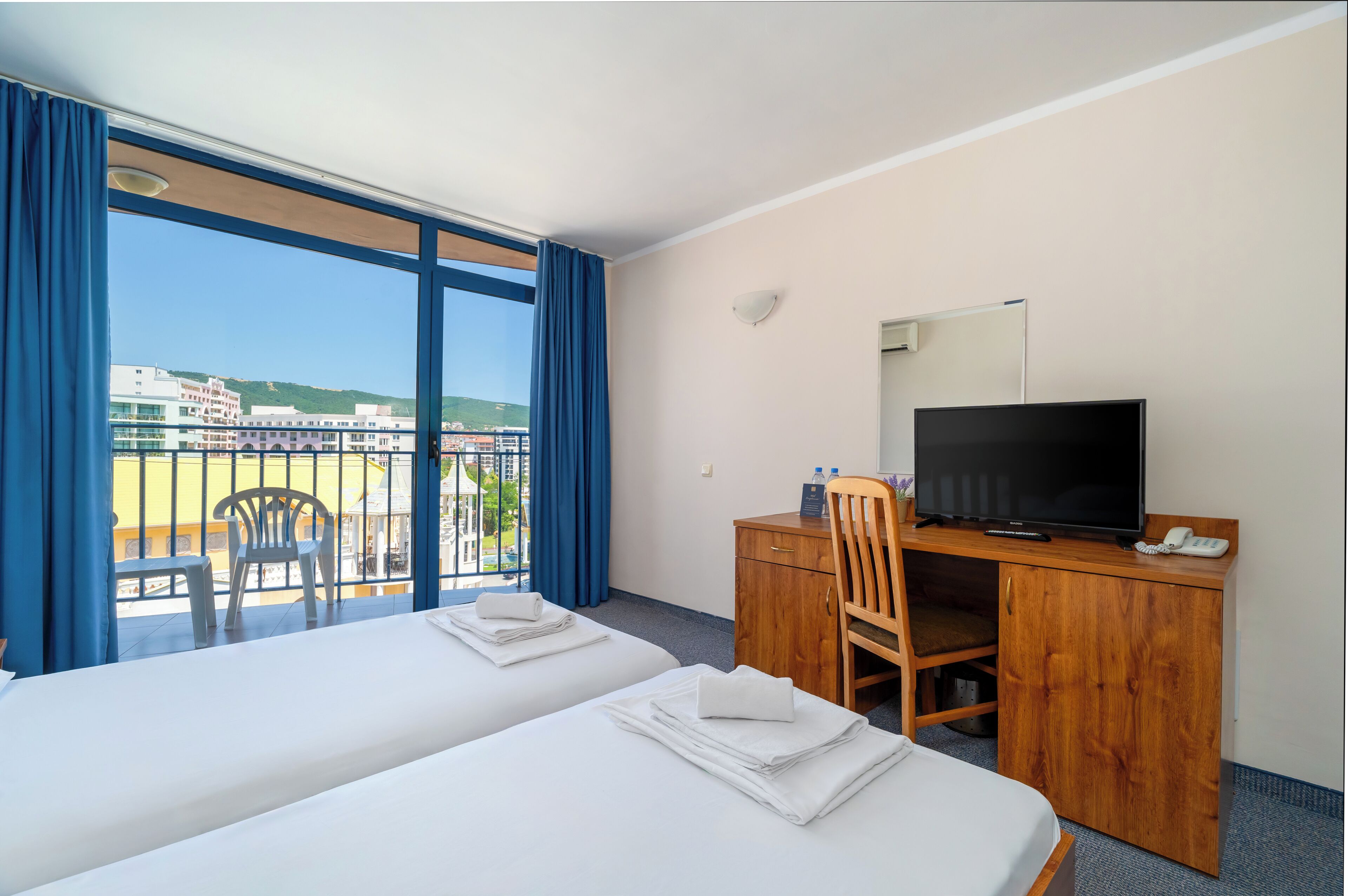 A hotel room with two beds, a desk with a television, a chair, and a balcony with a view of buildings and greenery.