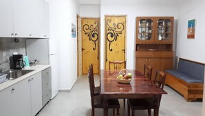 Une cuisine compacte avec des meubles blancs, une machine à café et une table à manger avec des chaises. On peut également apercevoir un meuble en bois avec des portes en verre et un banc bleu avec un coussin.