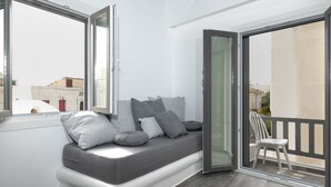 Een moderne woonkamer met een grijze bank, meerdere kussens en een glazen deur naar een buitenbalkon met een witte stoel.