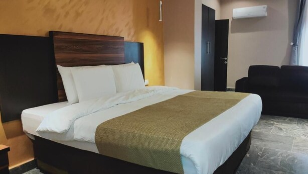 A hotel room with a large bed, a dark headboard, a brown throw blanket, and a black sofa. - En Apartment (Abuja)