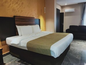 A hotel room with a large bed, a dark headboard, a brown throw blanket, and a black sofa. - En Apartment (Abuja)