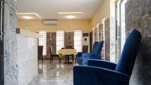 A dining area with a marble floor, blue chairs, a table with a yellow tablecloth, and a window with blinds. - En Apartment (Abuja)