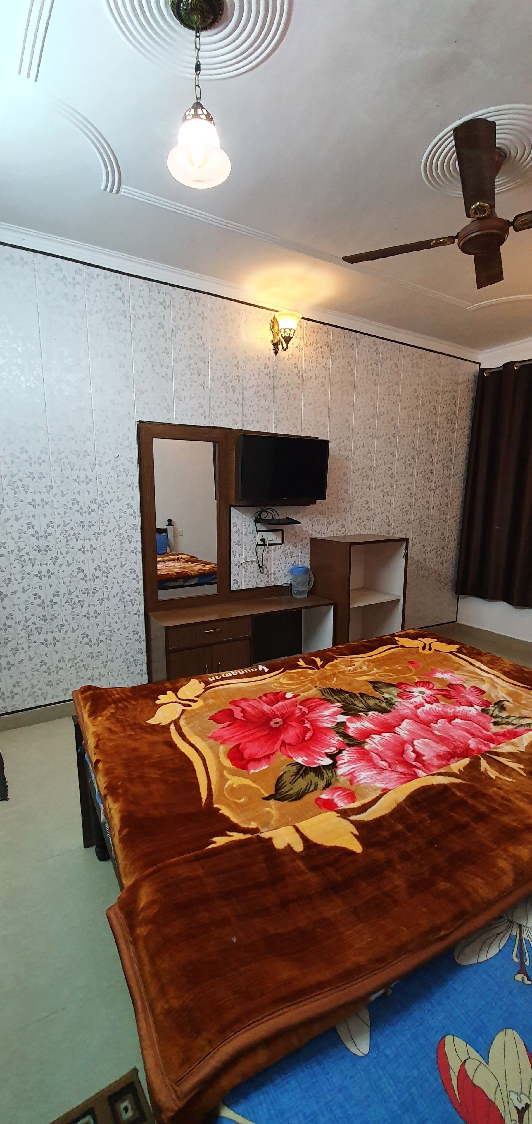 A hotel room with a bed, a television, a ceiling fan, and a wall-mounted light fixture.