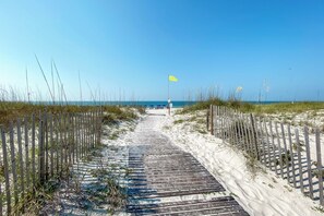 A wooden pathway leading to a beach with a yellow flag, surrounded by sand and grass.