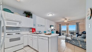 Una cocina moderna con armarios blancos, horno empotrado y lavavajillas. Hay una zona de comedor con un sofá azul y una mesa pequeña.