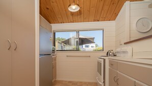 A compact kitchen with wooden ceiling, a window, and a view of a house. - Champion Bach - Pauanui Holiday Home (Pauanui)