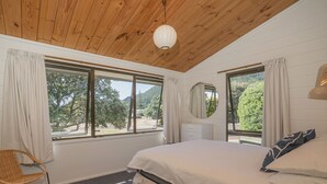 A bedroom with a wooden ceiling, a bed with white bedding, a round mirror, a dresser, and a chair. - Champion Bach - Pauanui Holiday Home (Pauanui)