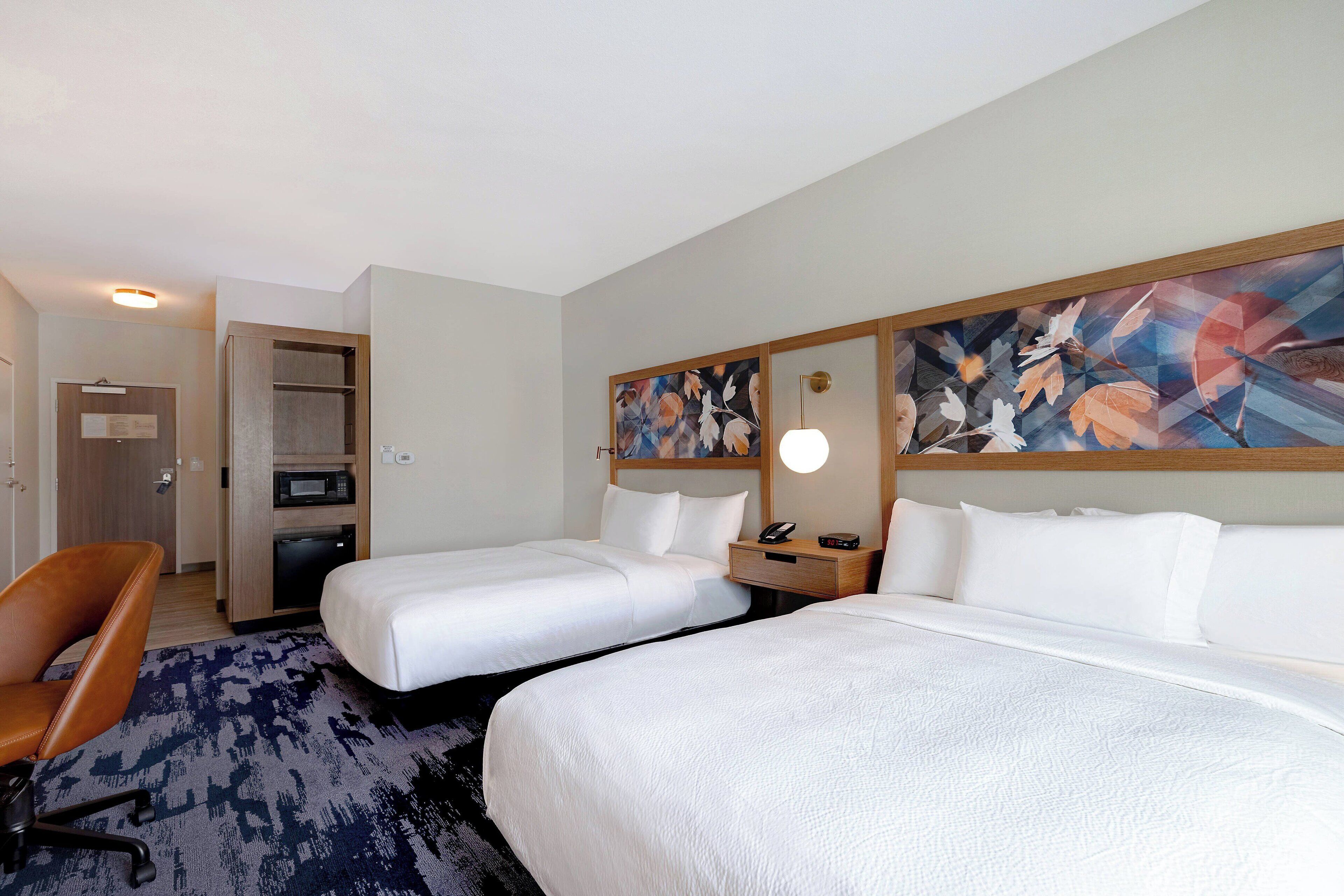 Quarto de hotel com duas camas de solteiro, uma escrivaninha com cadeira, micro-ondas e um quadro na parede.