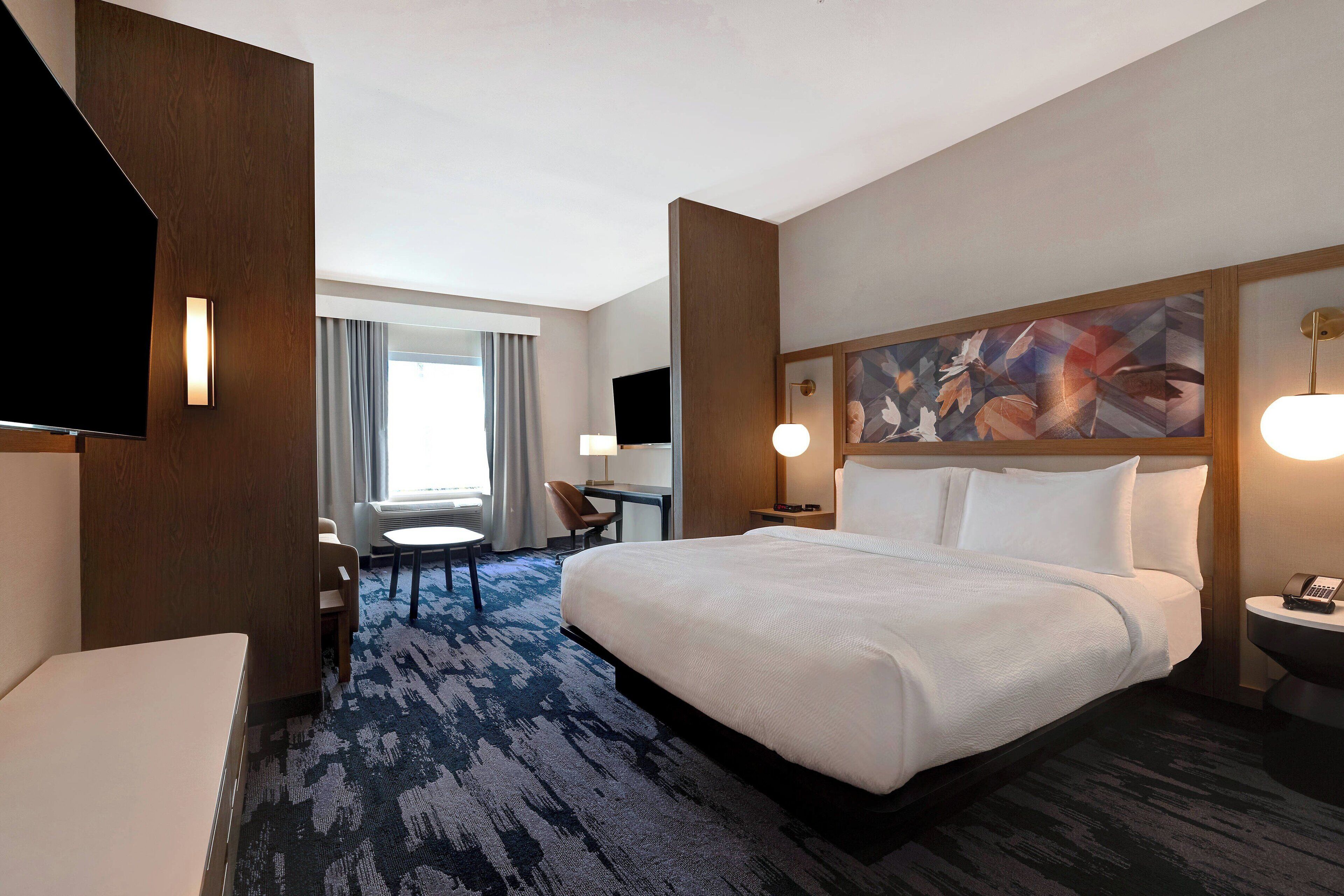 Habitación de hotel con una cama grande, un escritorio, una silla, un televisor y un cuadro en la pared.