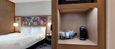 A hotel room with a bed, a nightstand, a lamp, a painting on the wall, a shelf with a microwave, a basket of toiletries, and a coffee maker.