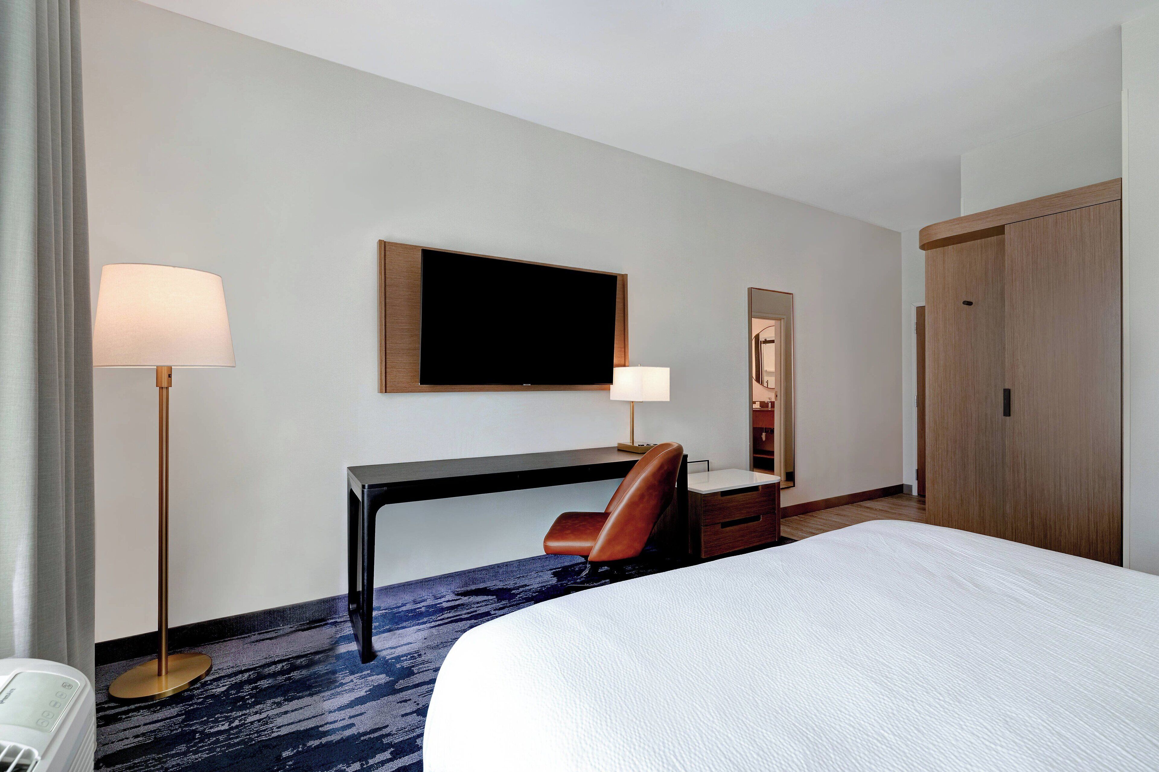 Habitación de hotel con cama, escritorio, silla, dos lámparas, un televisor de pantalla plana y un armario.