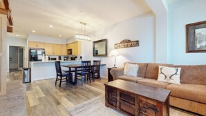 A spacious living area with a wooden coffee table, a sofa with decorative pillows, and a dining area with a wooden table and chairs. A kitchen with wooden cabinets and a large island is visible in the background. - On Sierra Star Golf Course, Washer/Dryer, Quiet Location & On-site Pool (Mammoth Lakes)