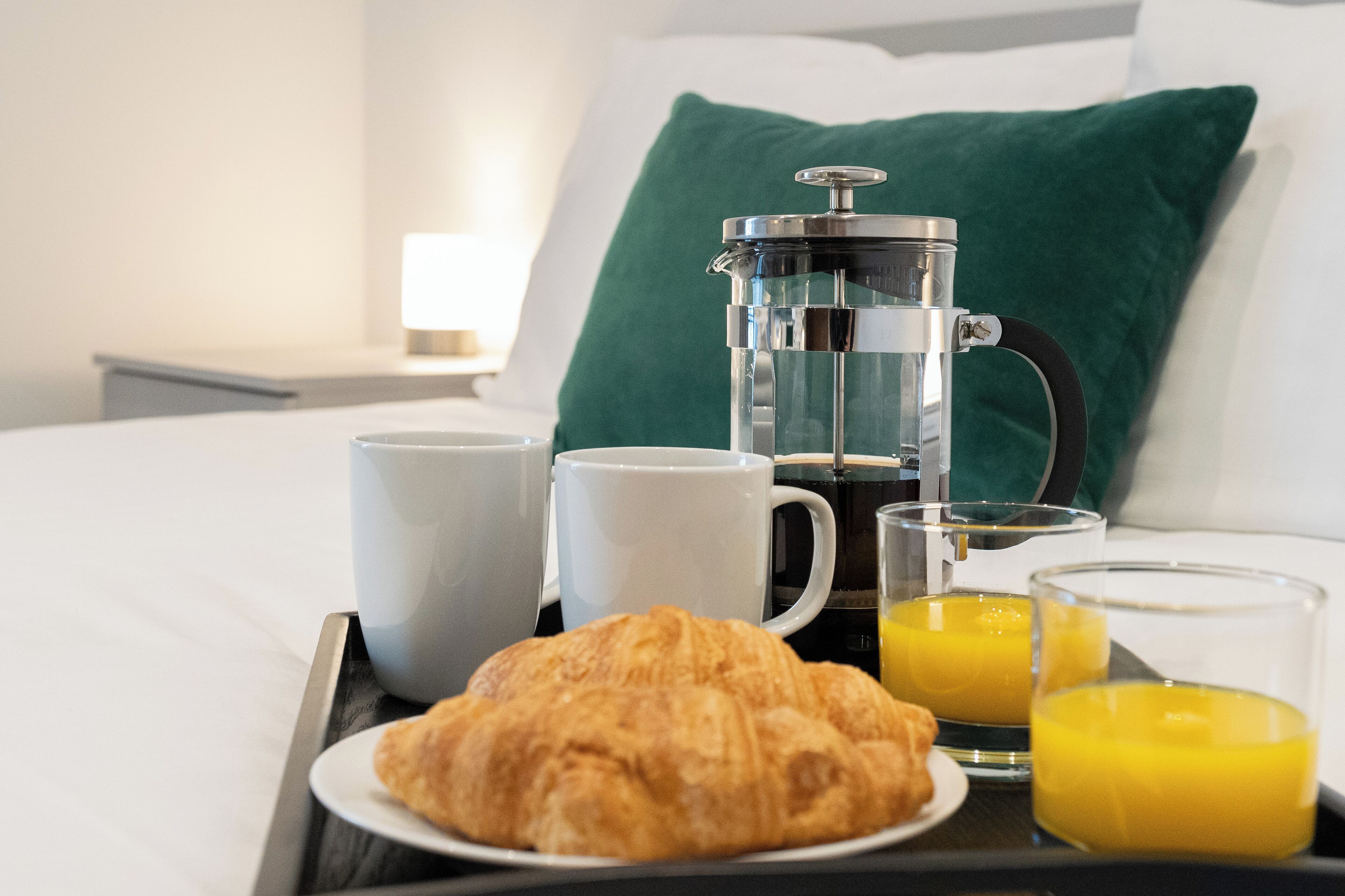 A breakfast tray with a croissant, two white mugs, a French press, and two glasses of orange juice on a bed with a green pillow and white bedding.