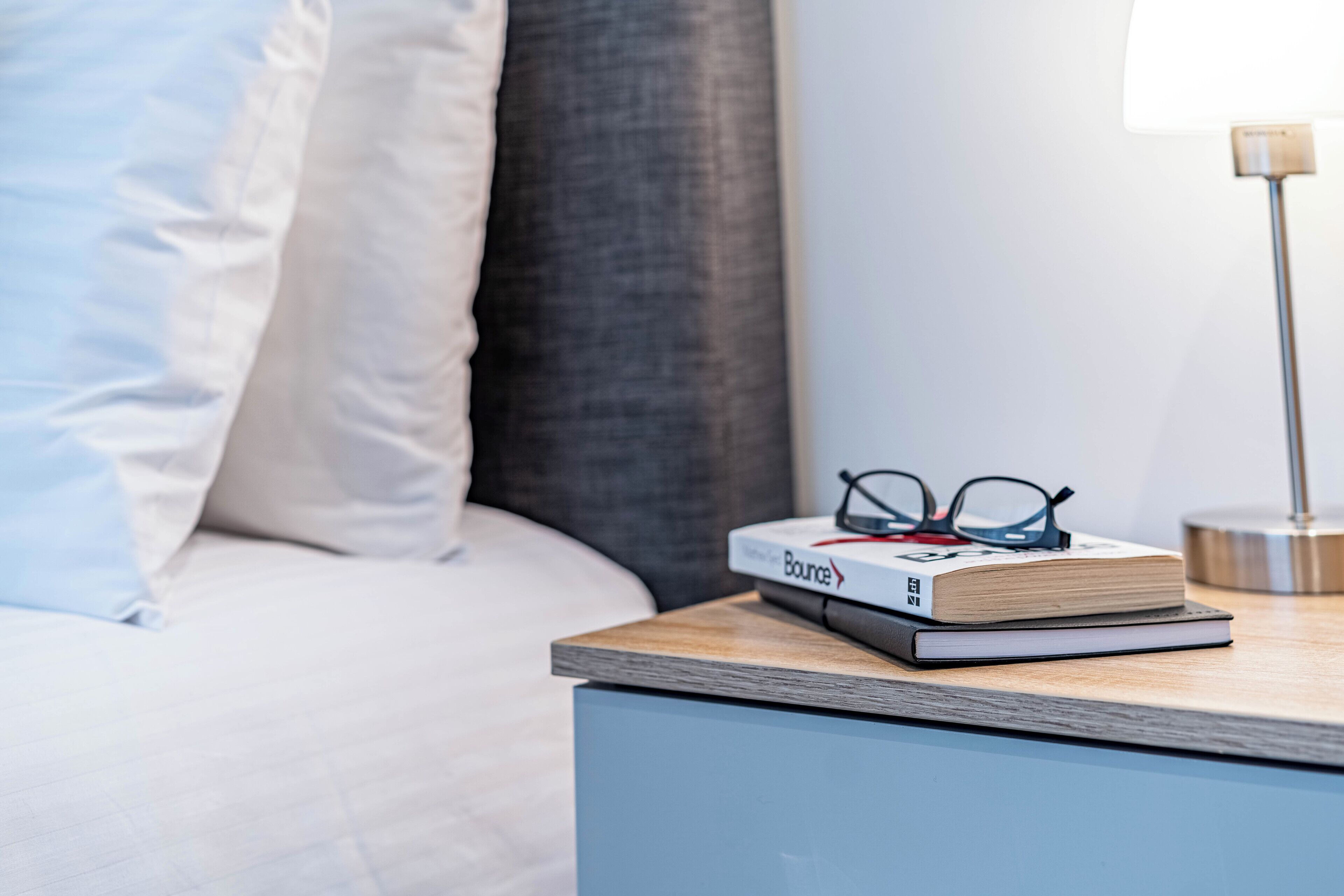 A neatly made bed with pillows, a bedside table with a book and glasses, and a lamp.