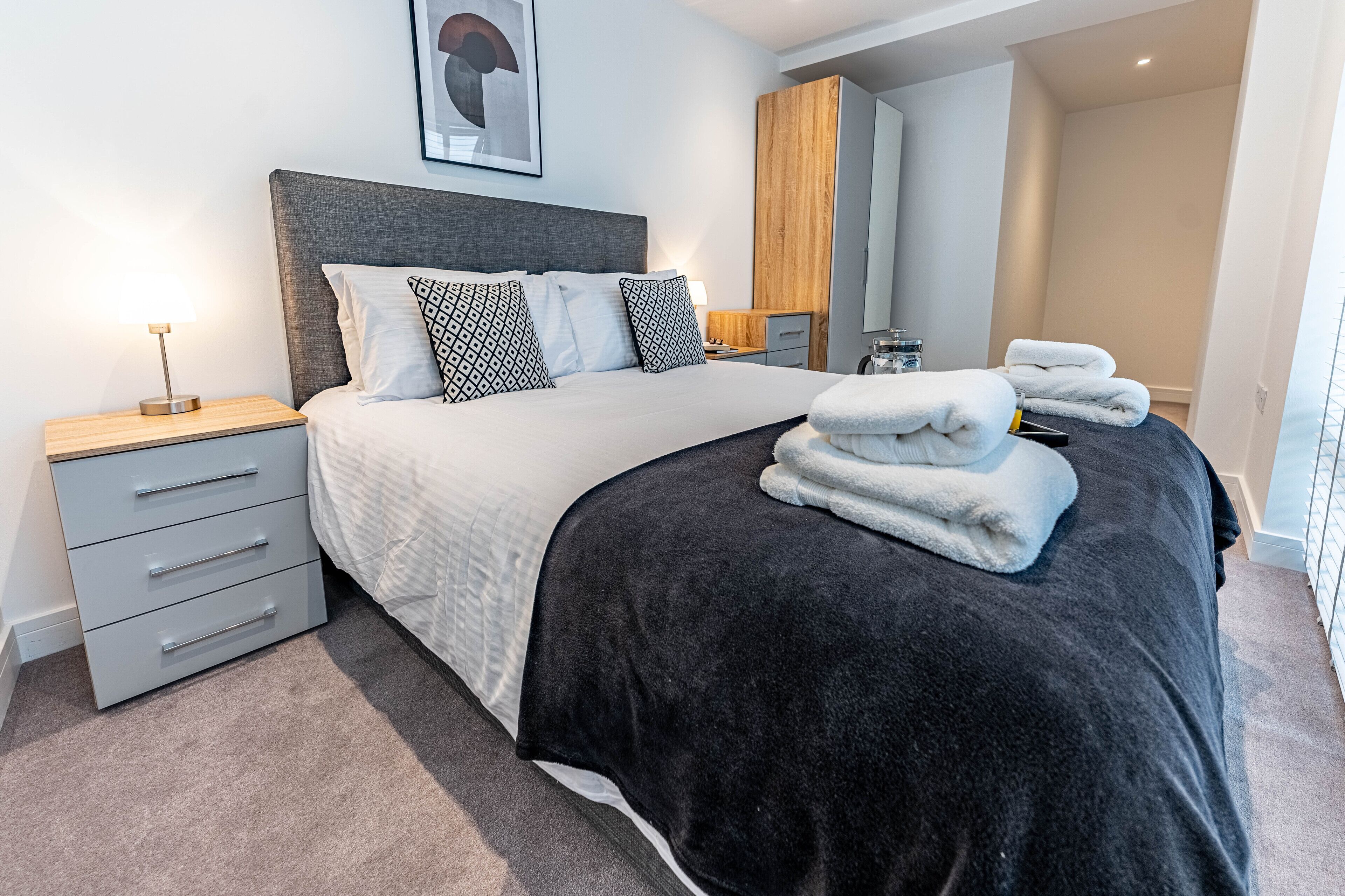 Una habitación de hotel moderna con una cama grande, una mesita de noche con lámpara, un armario y un baño visible a través de una puerta abierta.