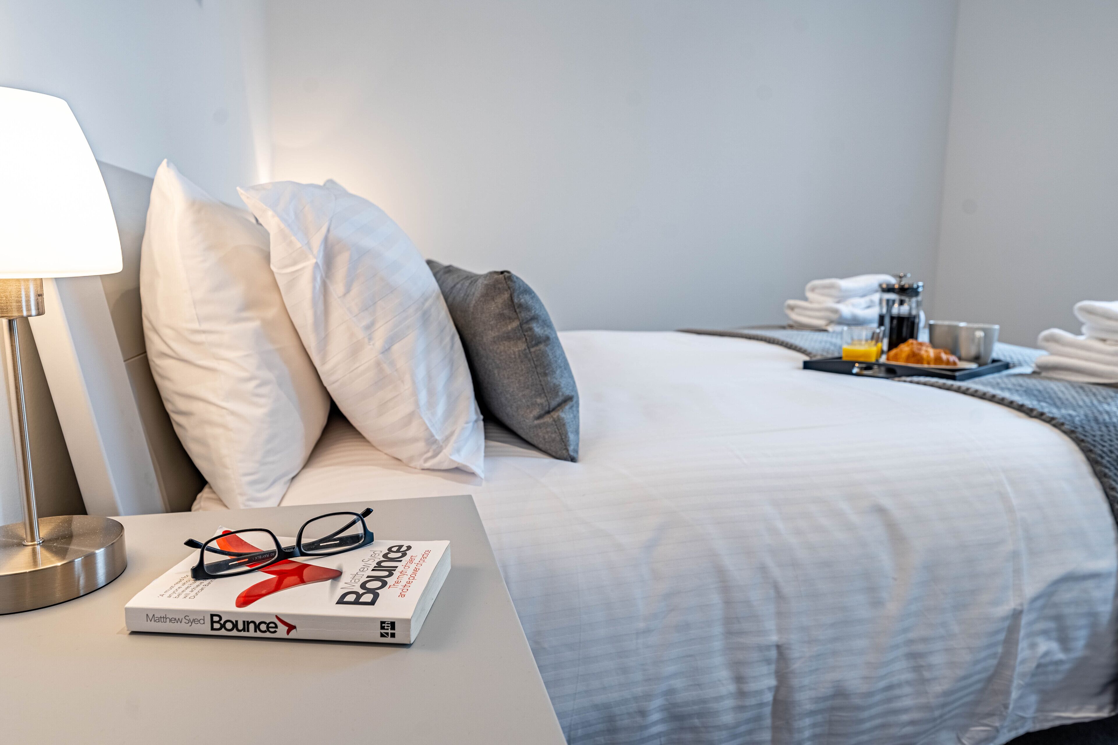 A neatly made bed with pillows, a bedside table with a book and glasses, and a tray with breakfast items.
