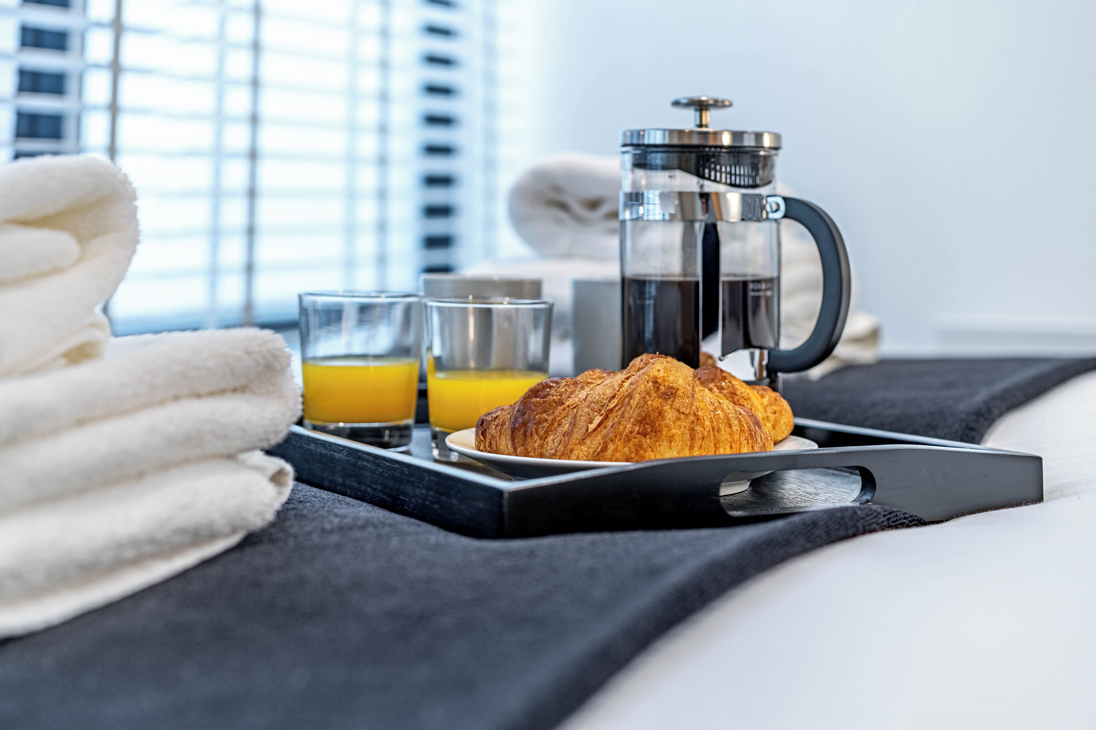 A breakfast tray with a croissant, two glasses of orange juice, and a French press coffee on a bed with a black tray.