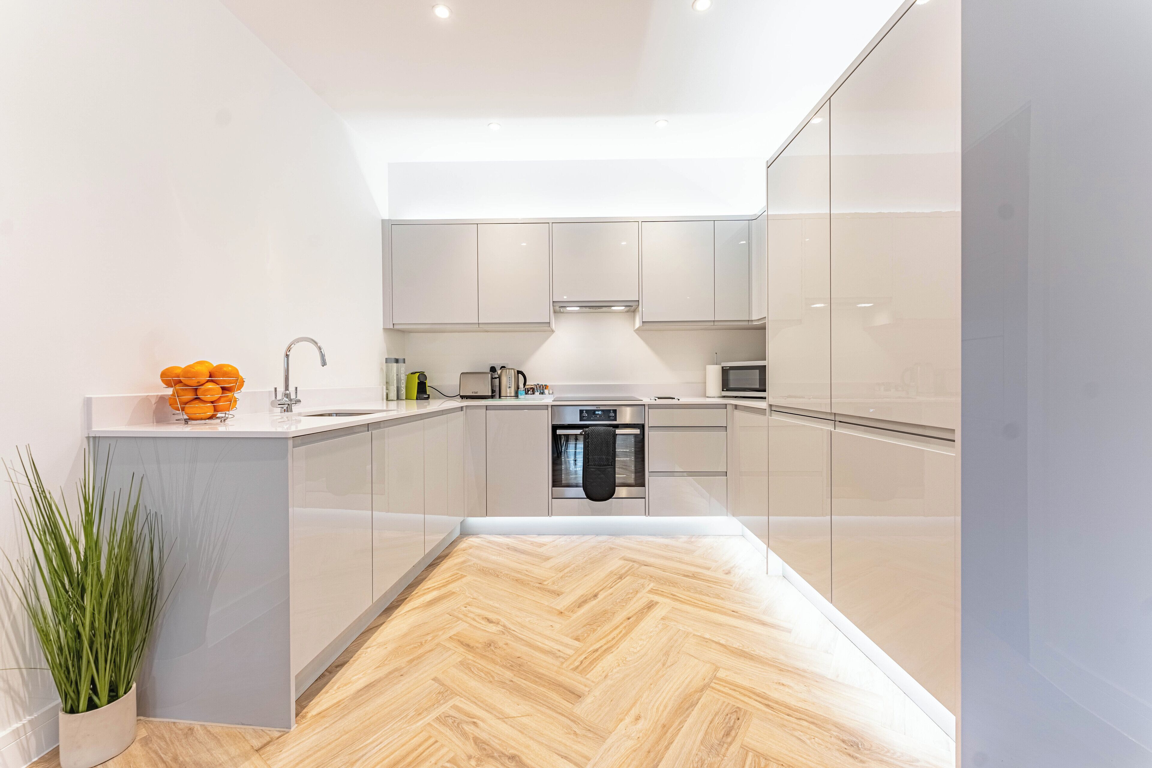 Uma cozinha moderna com piso de madeira em padrão herringbone, eletrodomésticos de aço inoxidável e armários brancos.