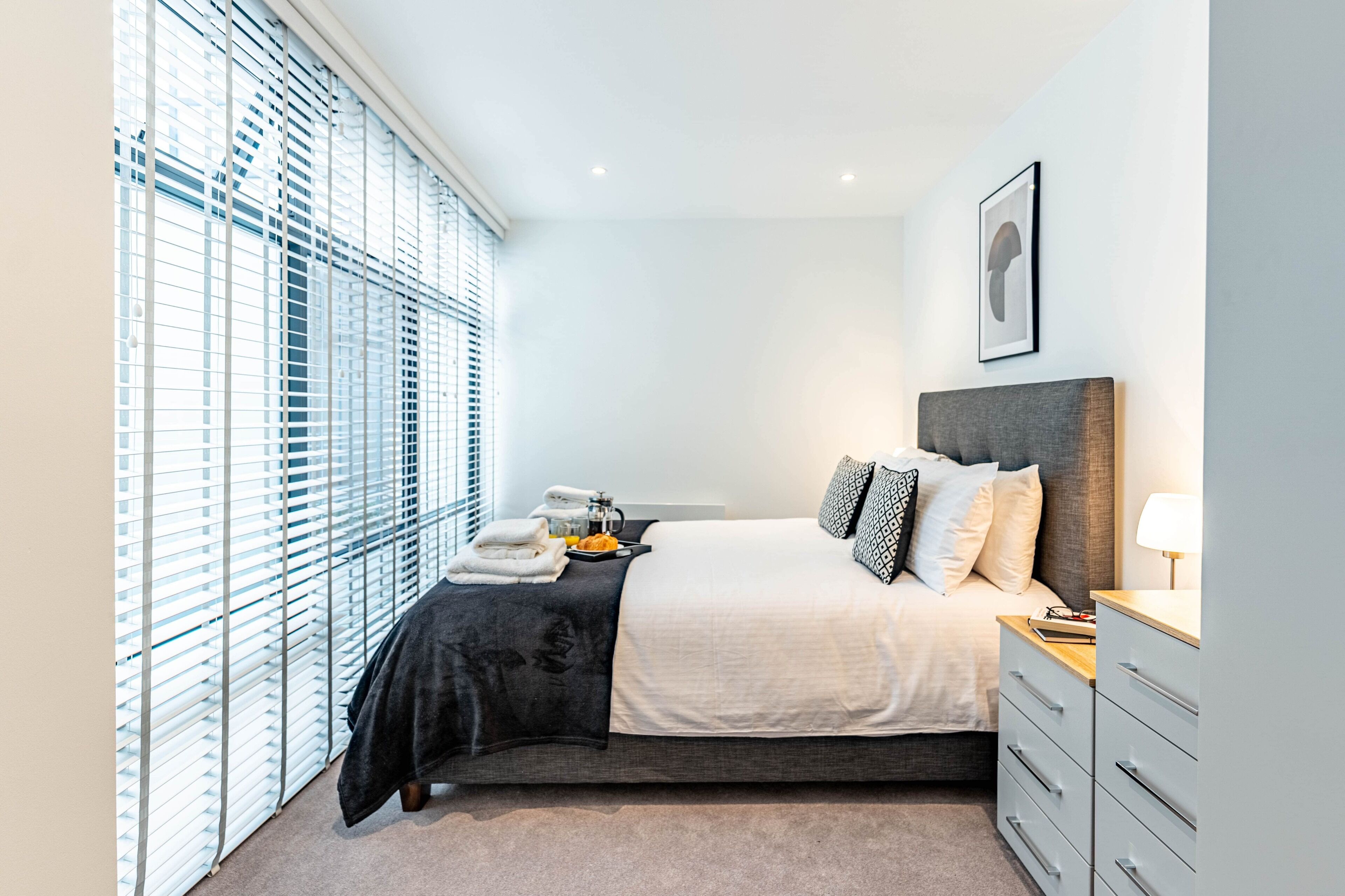 Een moderne slaapkamer met een groot bed, een nachtkastje, een lamp en een raam met jaloezieën.