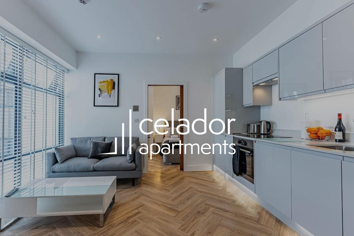 Un apartamento moderno con un sofá gris, una mesa de centro de vidrio, una cocina con armarios grises y un ventanal con persianas.