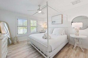 A white four-poster bed with a ceiling fan, a round mirror, and a bedside table with a lamp.