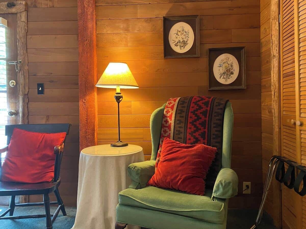 Ein gemütliches Zimmer mit einem grünen Sessel, einem roten Kissen, einem kleinen runden Tisch mit Lampe und Holzwänden.