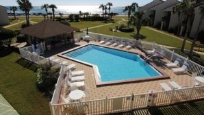 Una piscina rectangular con solárium azulejado, rodeada por una cerca blanca. Se observan sillas de descanso y un pequeño pabellón.