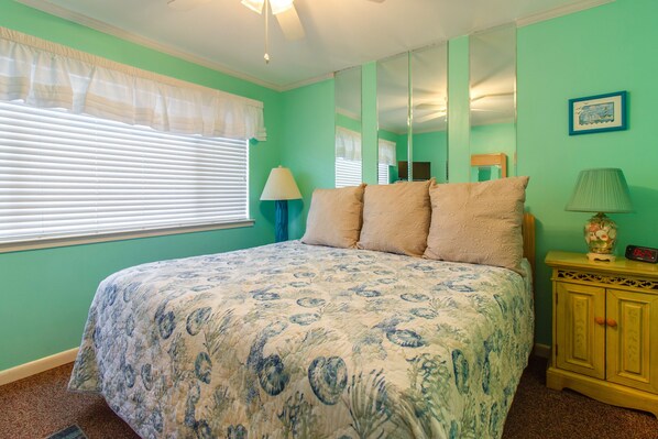 A bedroom with a bed, a nightstand, a lamp, a mirror, and a window with blinds. - Blue Surf 14A- Steps to the beach (Miramar)