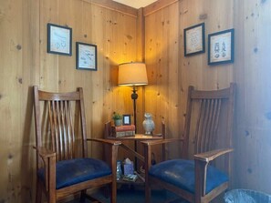 A corner room with wooden walls, two wooden chairs with blue cushions, a small table with a lamp, and framed drawings on the wall.