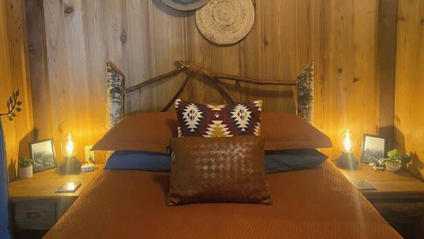 Quarto de madeira com uma cama, duas mesas de cabeceira e uma luminária fixada na parede.