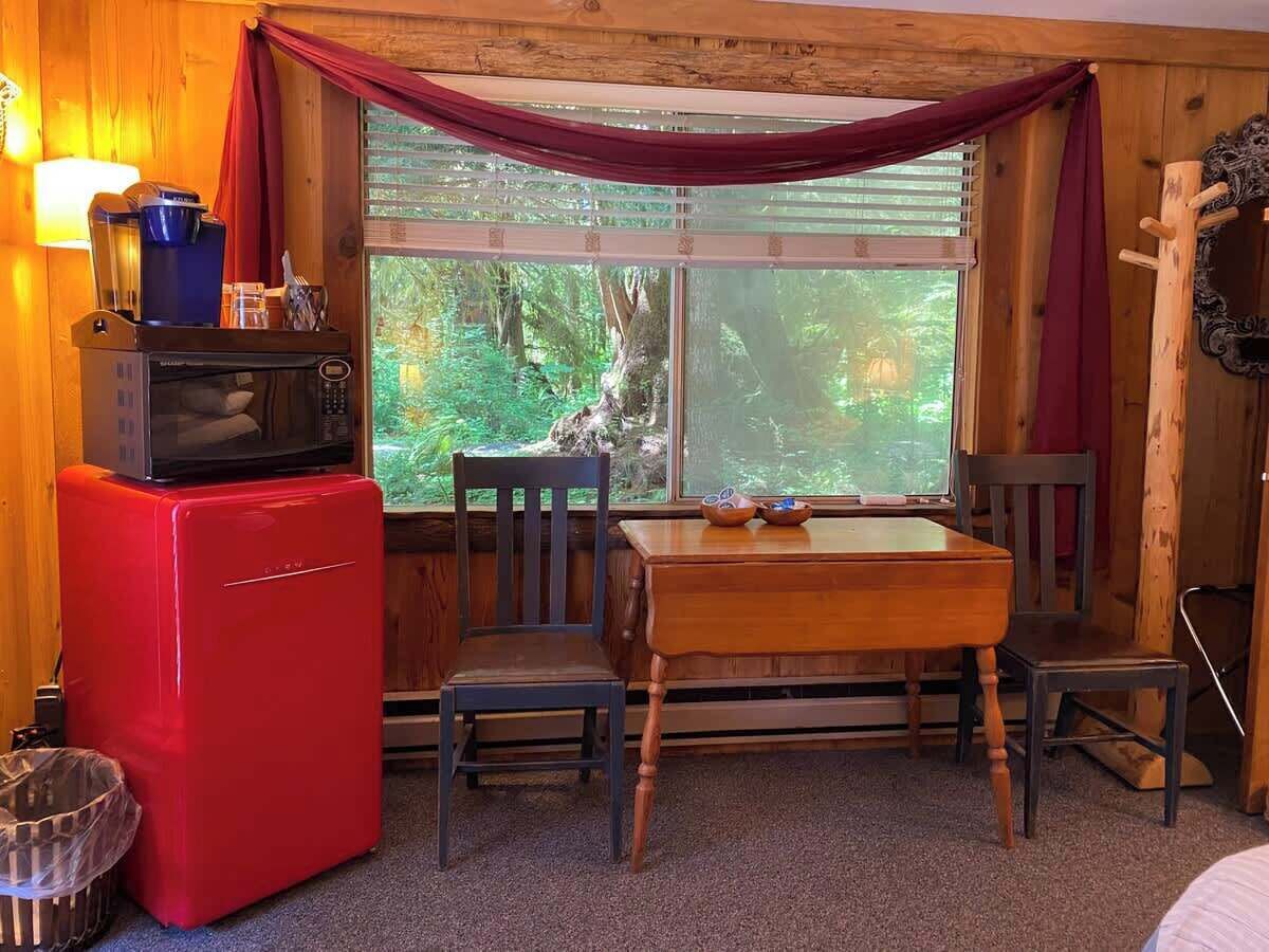 Ein kleiner Raum mit einem roten Kühlschrank, einem Holztisch und Stühlen. Ein Fenster mit Vorhängen bietet Blick ins Grüne.