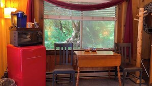 Quarto compacto com geladeira vermelha, mesa de madeira e cadeiras. Uma janela com cortinas oferece vista para o verde lá fora.