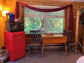 A compact room with a red refrigerator, wooden table, and chairs. A window with curtains offers a view of greenery outside.