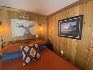 A wooden-paneled room with a bed, a wall-mounted sign indicating hiking trails, and a framed picture of a coastal landscape.