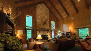Amplia sala de estar con chimenea de piedra, vigas de madera y ventanas grandes que ofrecen vistas al exterior.