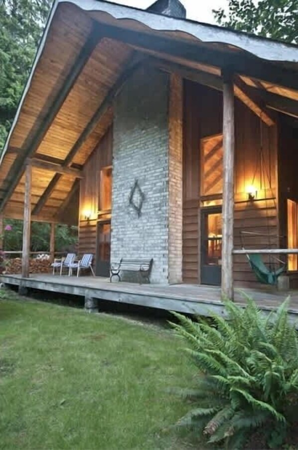 A wooden cabin with a stone wall, a covered porch with chairs, and a hammock.