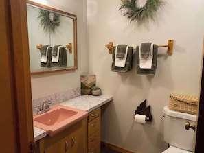 Une salle de bain avec un lavabo rose, des meubles en bois, un miroir, des serviettes accrochées, des toilettes et une couronne décorative.