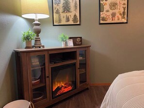 A room with a wooden cabinet containing a fireplace, a lamp, and framed botanical prints on the wall.