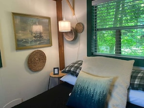 Une chambre à coucher avec un lit, un tableau encadré, une lampe et une vue sur la verdure par la fenêtre.