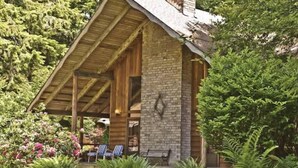 A rustic cabin with a wooden porch and brick walls, surrounded by lush greenery.