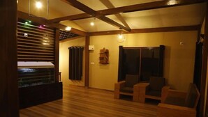 A spacious room with wooden flooring, a large window with blinds, and a seating area with wooden furniture. - Rustic Charm Wayanad - Barefoot Bungalow (Appapara)