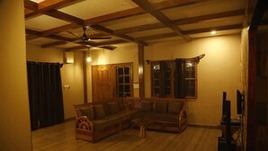 A living room with a wooden ceiling, a ceiling fan, a sectional sofa, a small coffee table, and a television. - Rustic Charm Wayanad - Barefoot Bungalow (Appapara)