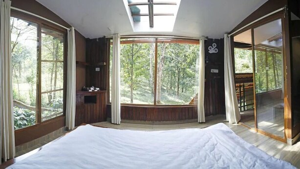 A bedroom with a large bed, a skylight, and a view of a forest. - Rustic Charm Wayanad - Barefoot Bungalow (Appapara)