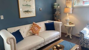 Una sala de estar con un sofá color beige, cojines azules, una ilustración enmarcada de peces, una mesita auxiliar con una lámpara y una silla estampada.