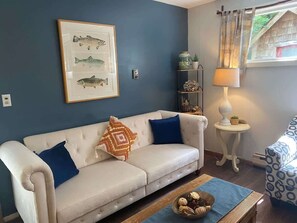 A living room with a beige sofa, blue cushions, a framed fish illustration, a small side table with a lamp, and a patterned chair.