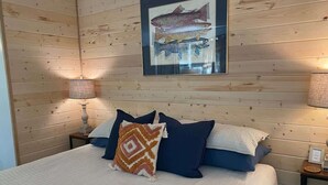 A wooden-paneled room with a bed, two bedside lamps, and a framed fish illustration on the wall.