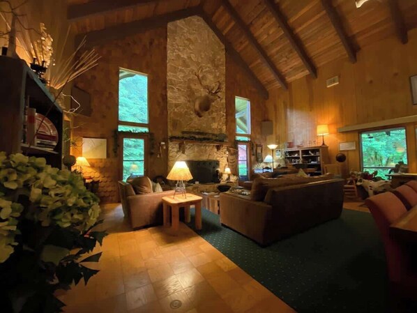 Amplia sala de estar con chimenea de piedra, vigas de madera y ventanas grandes que ofrecen vistas al exterior.