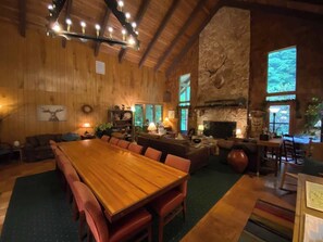 A spacious room with a large wooden dining table, comfortable seating, a fireplace, and large windows allowing natural light.